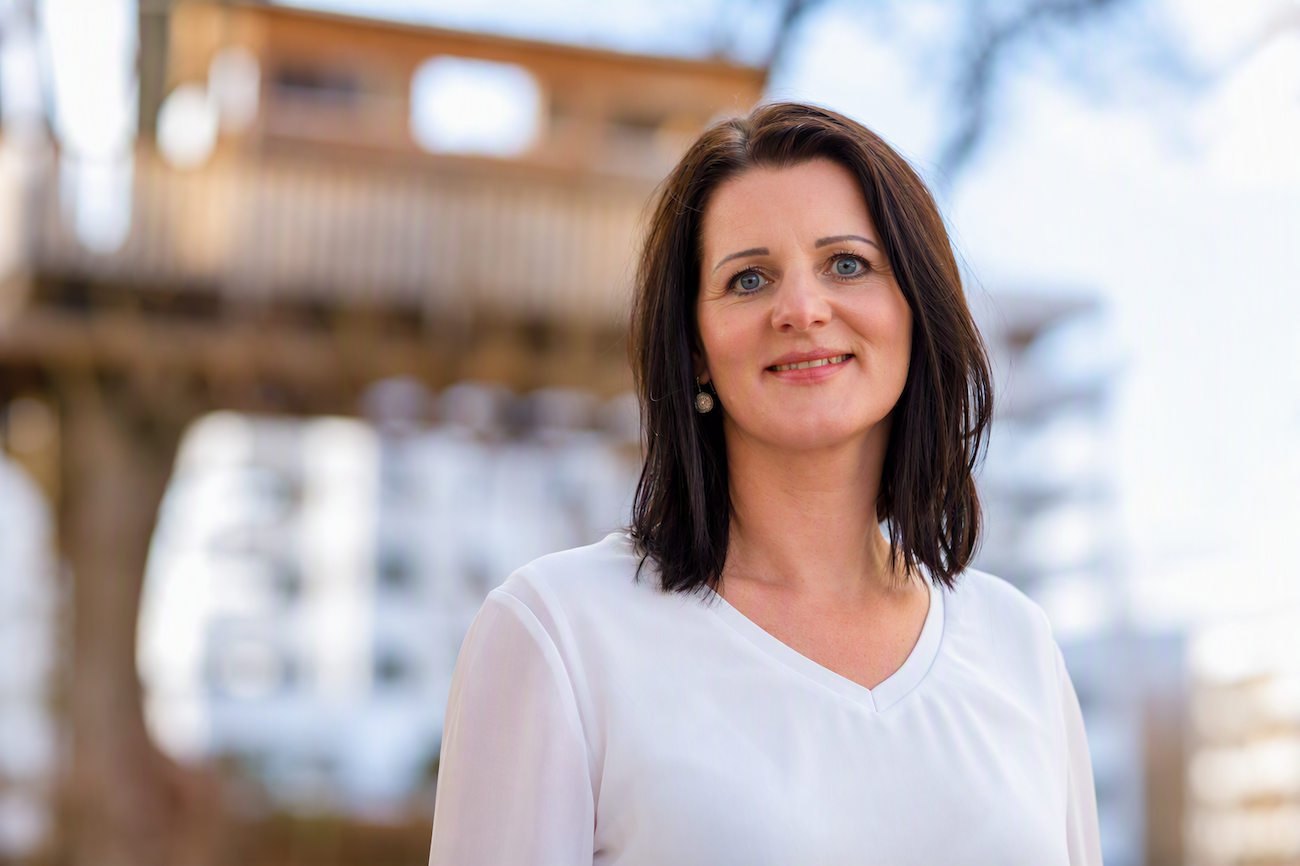 Frau mit dunklem Haar in weißer Bluse lächelnd im Freien, unscharfer Hintergrund.