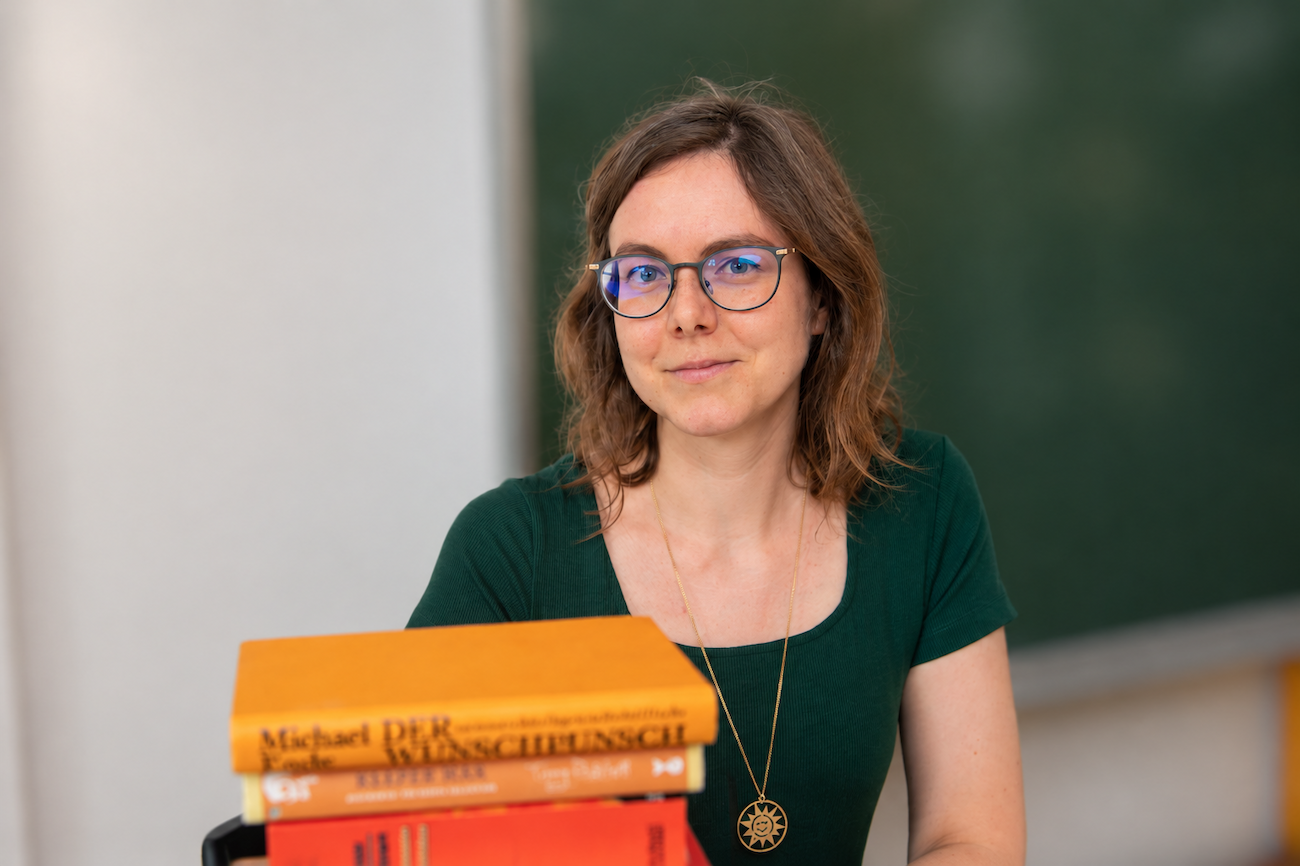 Eine Frau mit Brille sitzt neben einem Stapel Bücher vor einer Kreidetafel.