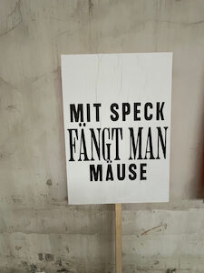 Ein Schild mit dem deutschen Text Mit Speck fängt man Mäuse an einer Wand.