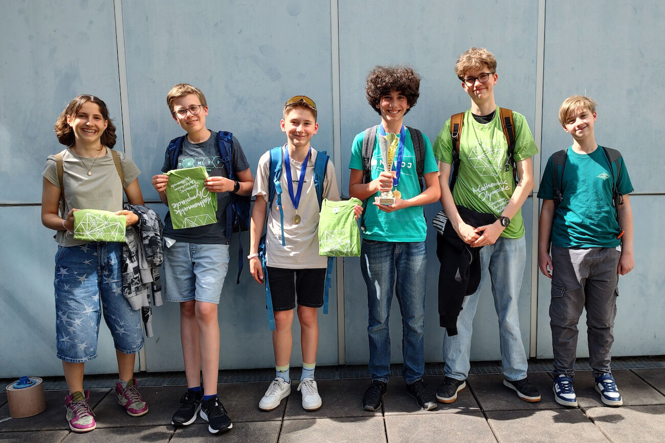 Sechs lächelnde Kinder stehen mit Medaillen und Trophäen vor einer hellblauen Wand.