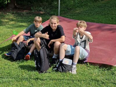 Drei Jungen sitzen mit Rucksäcken im Gras, einer zeigt den Daumen nach oben und lächelt.