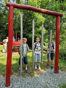 Drei Jungen spielen auf einem Glockenspiel im Freien, eingerahmt von roten Pfosten in einem Park.