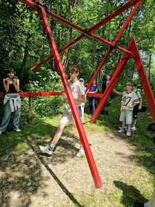 Eine Gruppe von Kindern steht um eine große rote Metallskulptur im Freien im Wald.