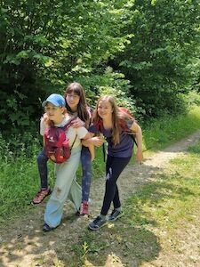 Drei Mädchen wandern; eine nimmt sie Huckepack, während sie auf einem Waldweg lächeln.