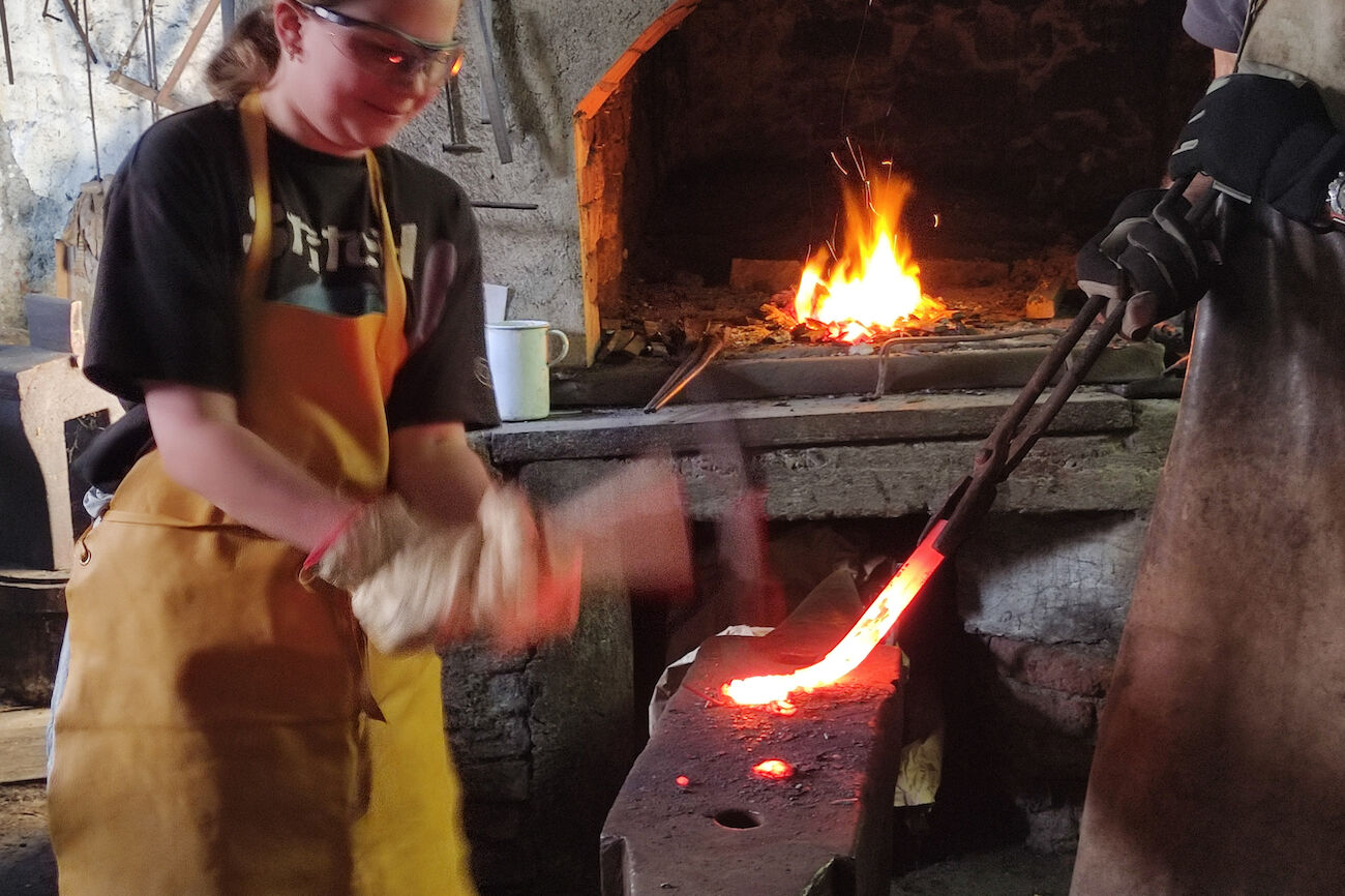 Eine Person schmiedet heißes Metall in einer Werkstatt, während im Hintergrund ein Feuer lodert.