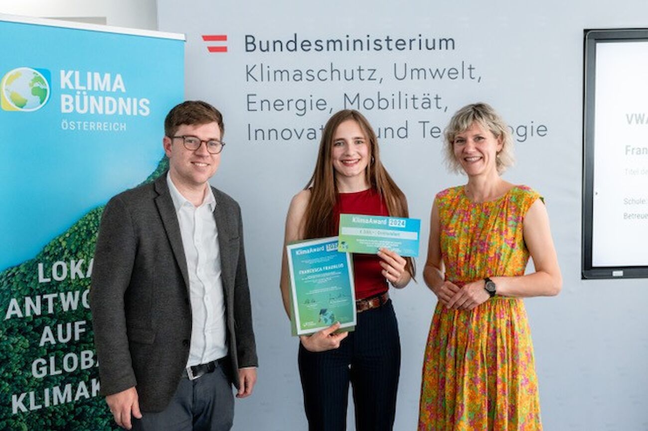 Drei Personen bei einer Preisverleihung, eine hält eine Urkunde und lächelt.